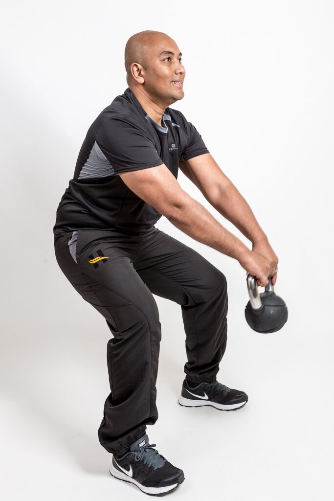 On voit une personne en train de faire un squat avec une kettlebell sur un fond blanc avec des vêtements noir et gris